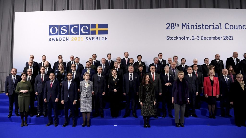 Många personer i kostym och dräkt på ett gruppfoto framför en vägg där det står: OSCE Sweden 2021.
